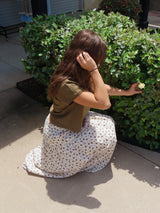 Person in a floral skirt and green top sitting on the ground next to a bush.